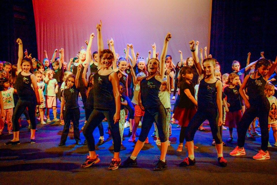 Young dancers dressed in black with their arms in the air. On the Amey Theatre stage with colourful lighting.